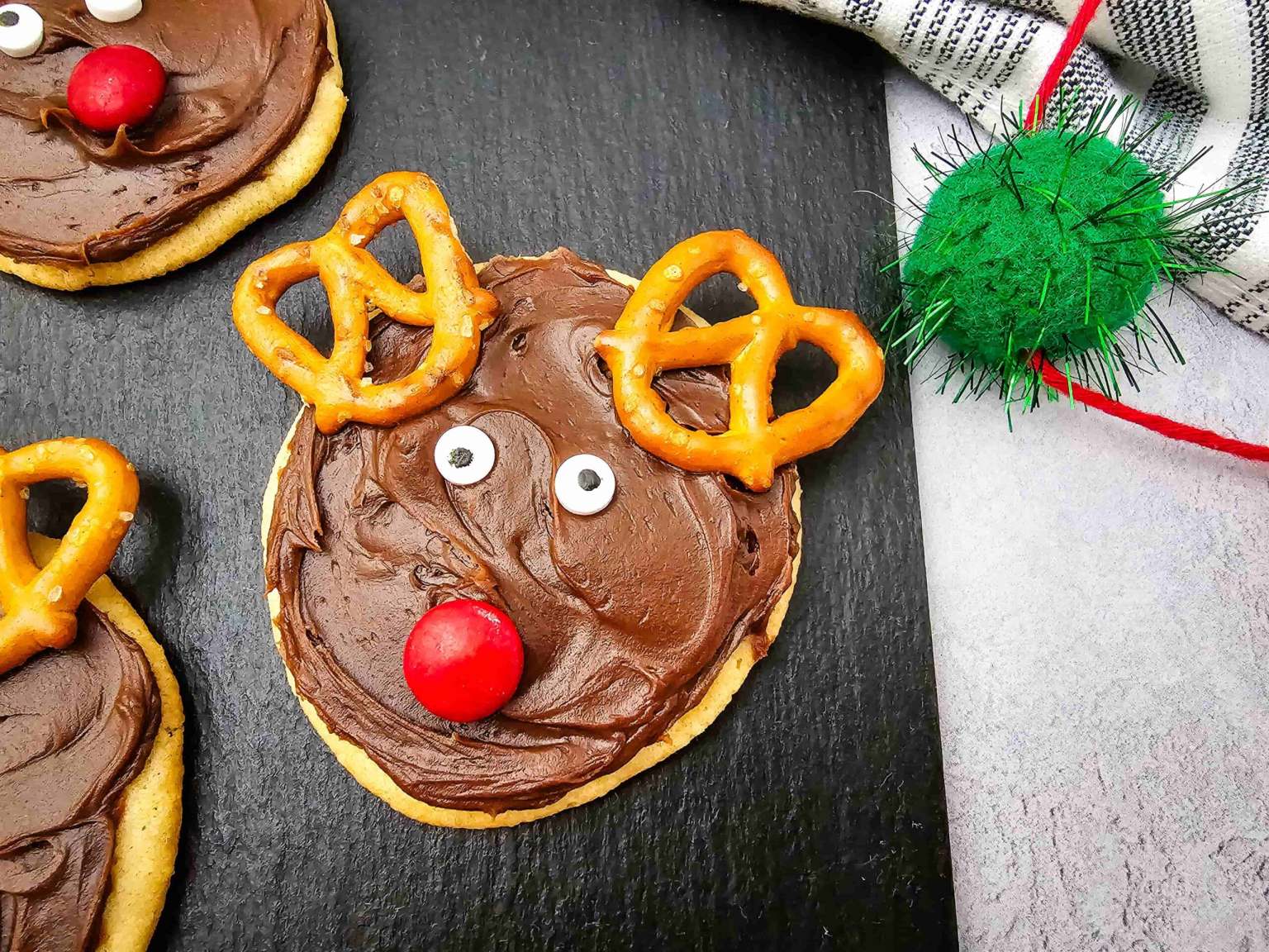 A cookie decorated with chocolate frosting, pretzel antlers, candy eyes, and a red candy nose to resemble a reindeer, placed on a dark surface next to a green ornament.