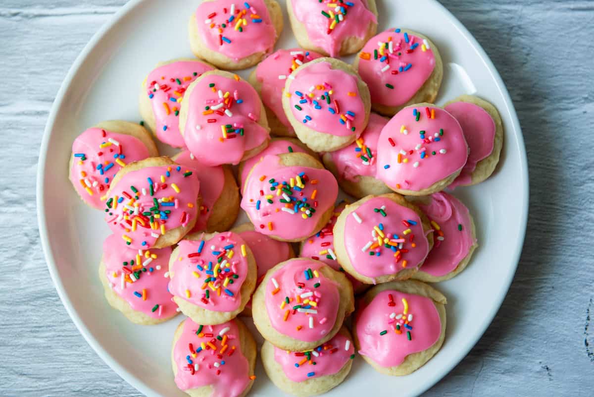 A white plate filled with round sugar cookies topped with pink icing and multicolored sprinkles, set on a light wooden surface.