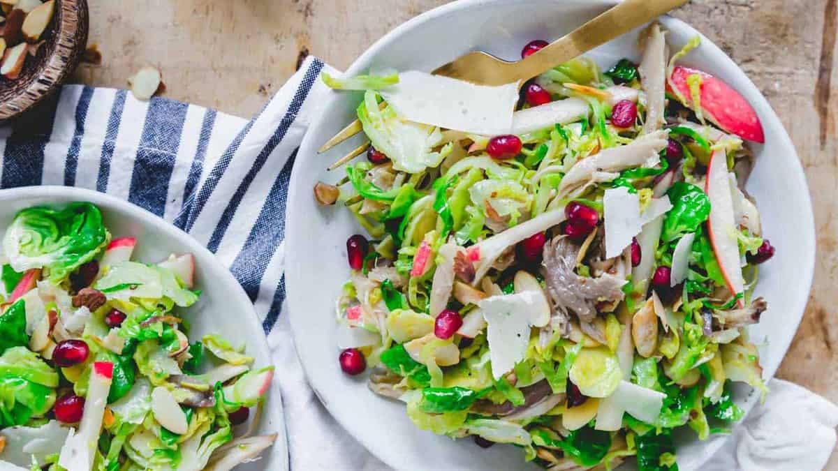 A bowl of salad with Brussels sprouts, apple slices, pomegranate seeds, shaved cheese, almonds, and shredded chicken, served with a fork on a striped napkin.