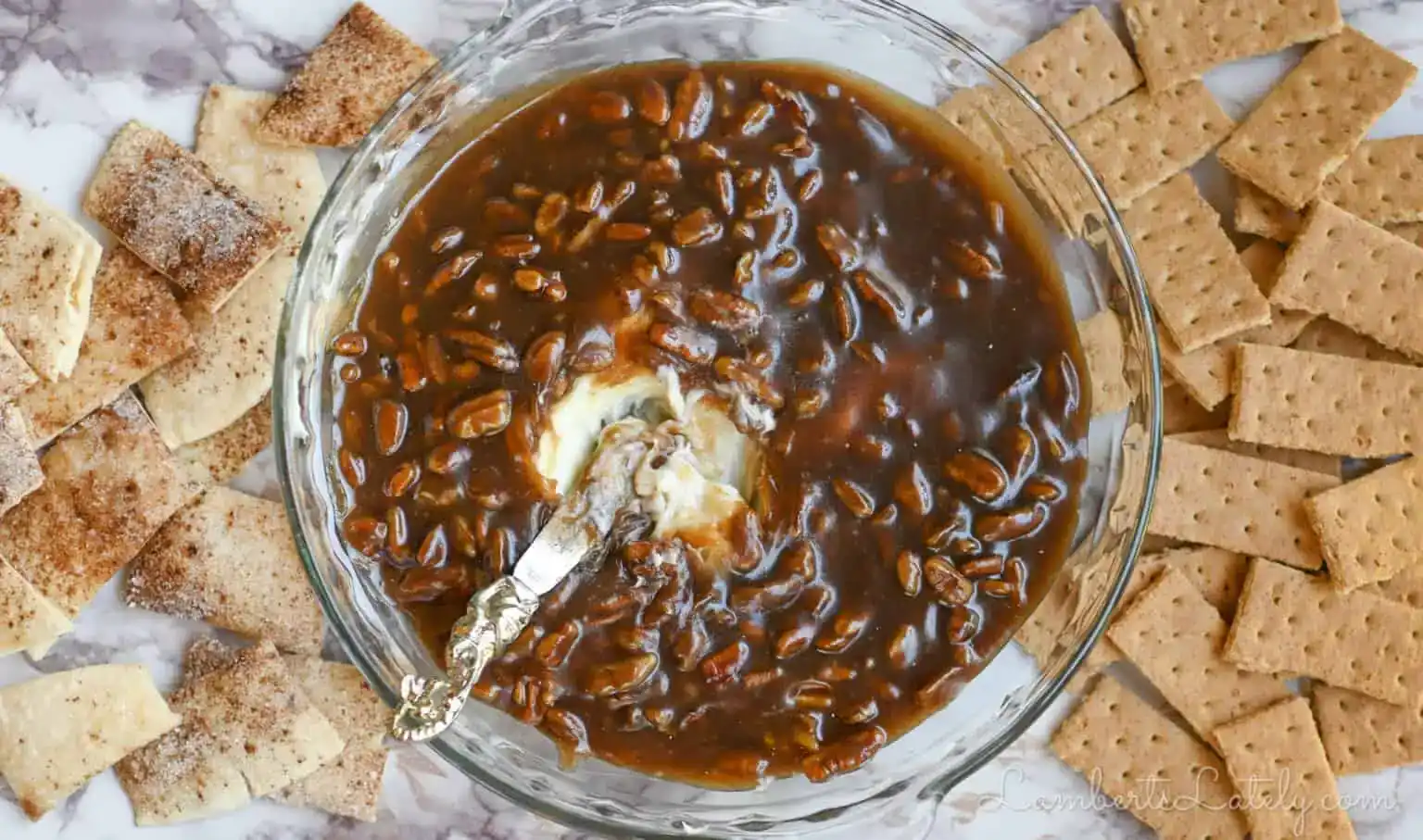 A glass bowl filled with a dark brown, gooey dip containing marshmallows and chocolate, surrounded by graham crackers and cinnamon sugar pita chips.