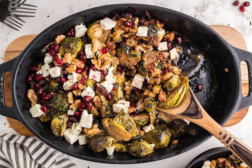 A black serving dish filled with roasted Brussels sprouts, feta cheese cubes, walnuts, and pomegranate seeds, with a wooden serving spoon beside the food.