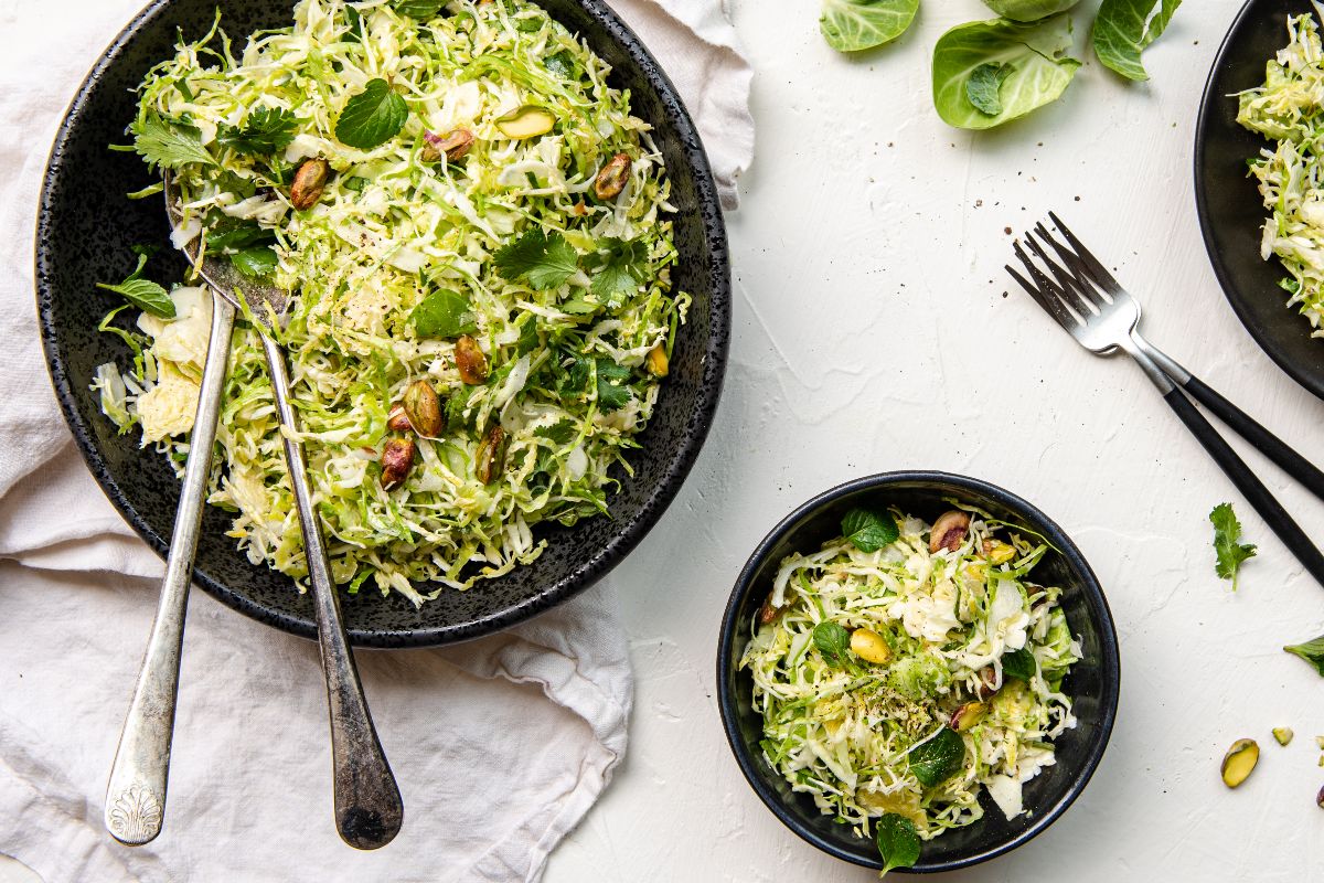 Two bowls of shredded Brussels sprout salad with herbs and pistachios sit on a white surface, next to forks and loose Brussels sprout leaves.