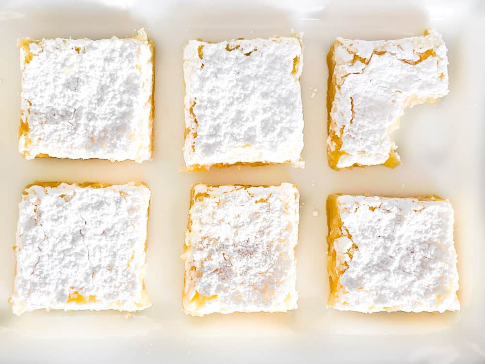 Six lemon bars topped with powdered sugar are arranged in two rows on a white surface; one bar has a bite taken out of it.