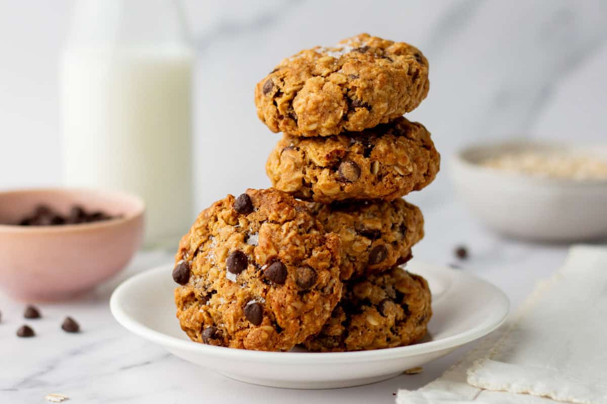 Four chocolate chip oatmeal cookies are stacked on a white plate, with a bottle of milk and bowls of chocolate chips and oats in the background.
