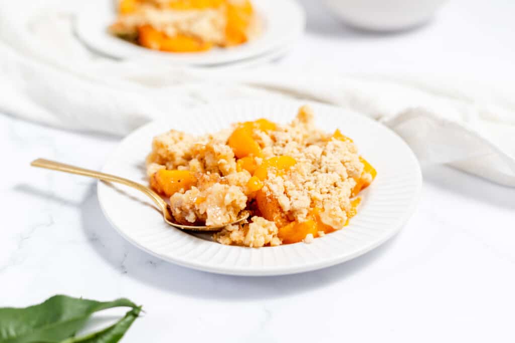A white plate with a serving of peach crumble and a spoon, set on a white surface with another plate in the background.