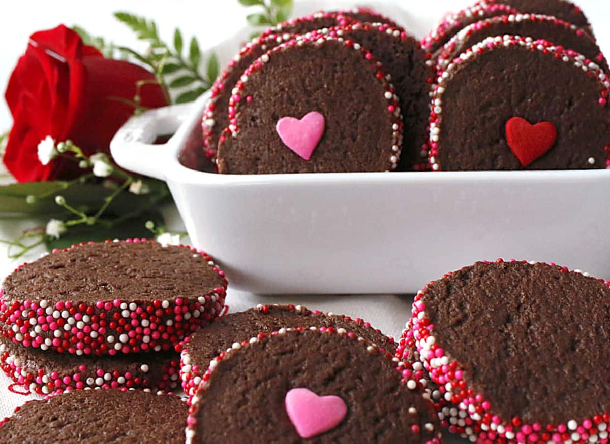 Chocolate cookies with heart-shaped decorations and red, white, and pink sprinkles arranged in and around a white dish; a red rose is in the background.