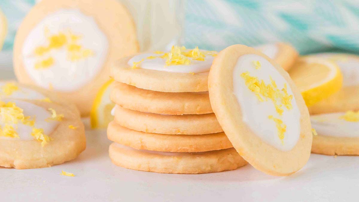 A stack of round sugar cookies with white icing and lemon zest on top, surrounded by more cookies on a light surface.
