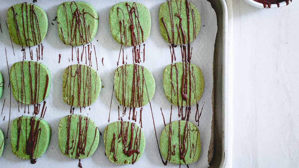 Rows of round green cookies on a parchment-lined baking sheet, drizzled with chocolate.