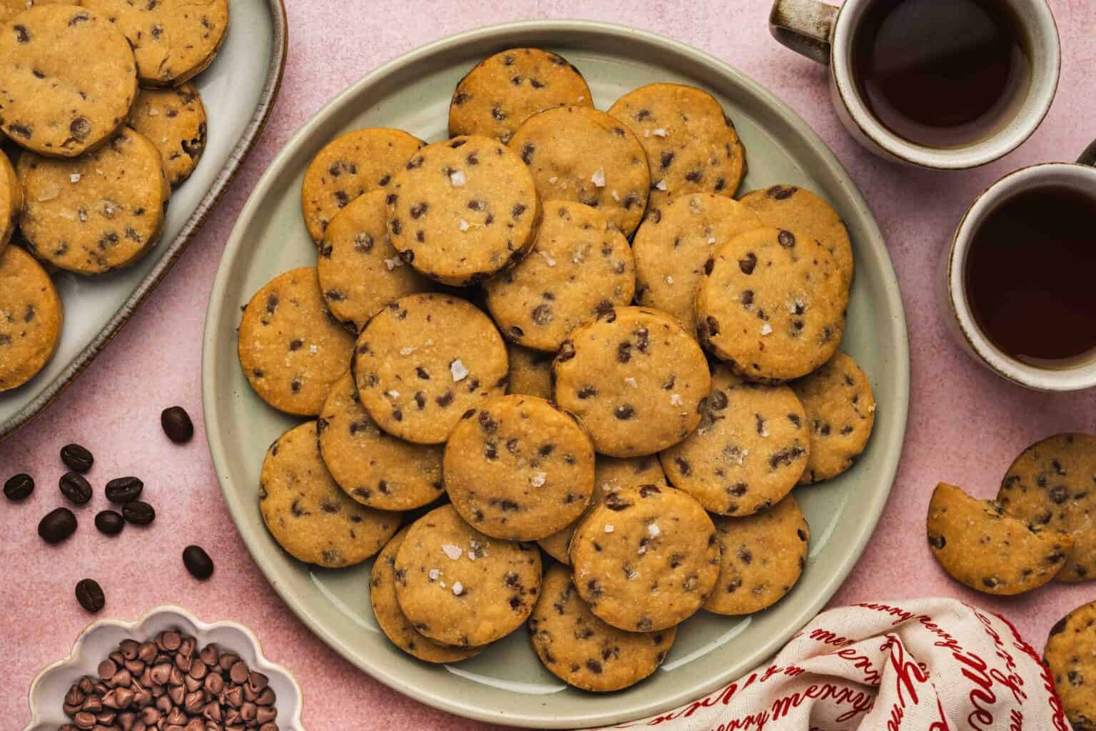A plate of chocolate chip cookies sprinkled with sea salt, surrounded by coffee cups, coffee beans, and a festive napkin on a pink surface.