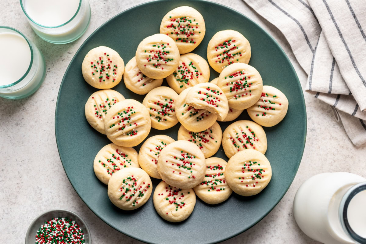 A green plate with round, flat cookies topped with red, green, and white sprinkles, surrounded by glasses of milk and a striped napkin.