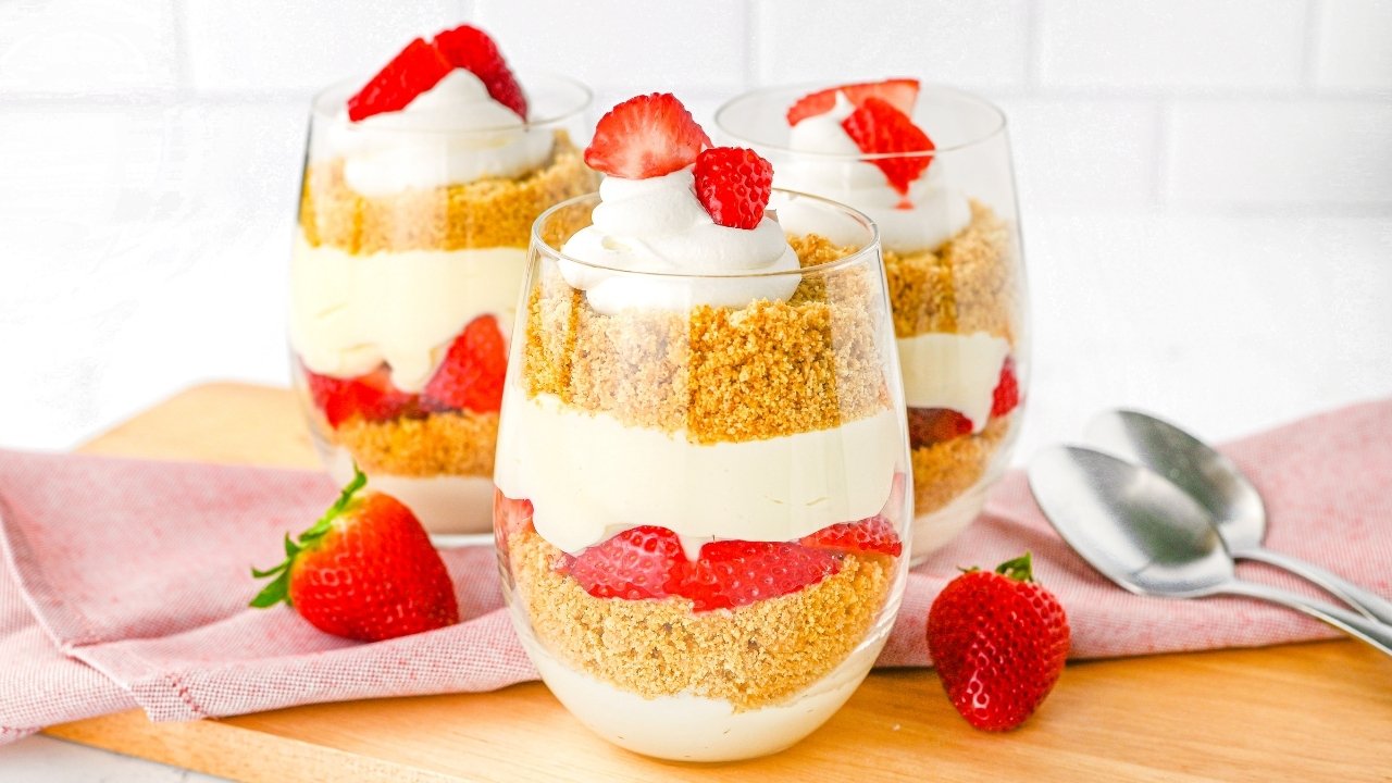 Three glass cups filled with layered cheesecake, graham cracker crumbs, whipped cream, and sliced strawberries, placed on a wooden board with two spoons and a fresh strawberry nearby.