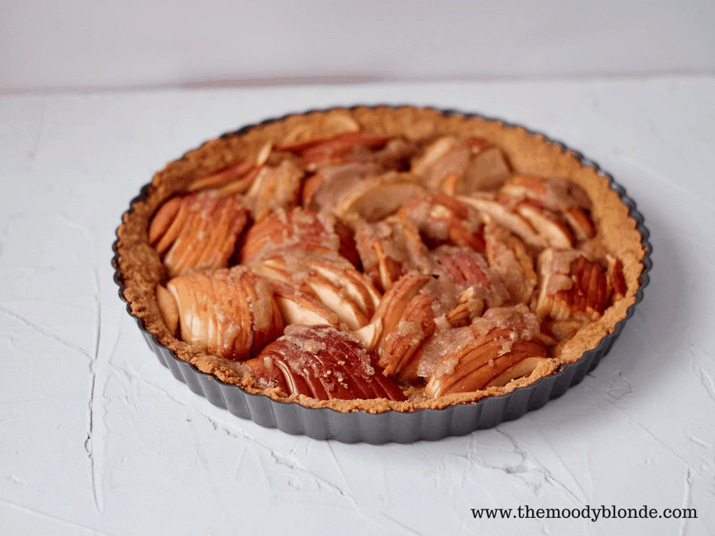 An apple tart with a golden-brown crust and sliced apples sits in a round baking tin on a white textured surface.