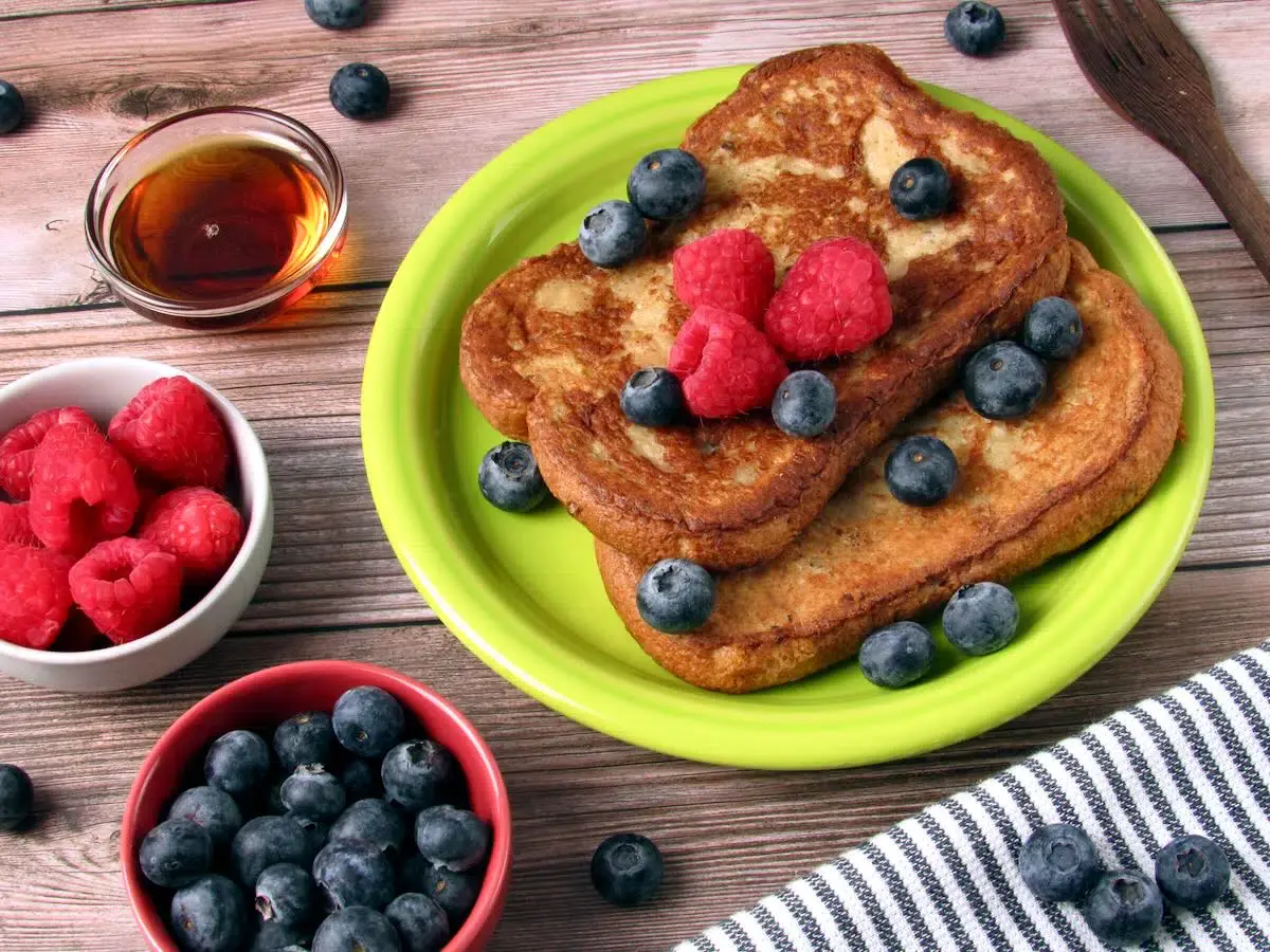 Two slices of French toast on a green plate, topped with raspberries and blueberries, with extra berries and syrup in bowls nearby on a wooden table.