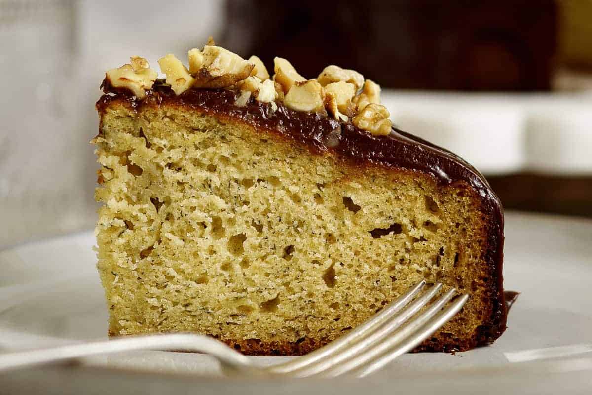 A slice of banana cake topped with chocolate frosting and chopped walnuts, served on a plate with a fork.