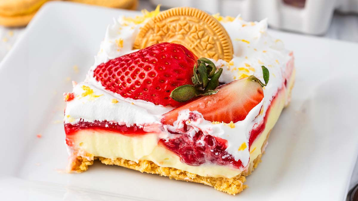 A dessert bar with a crumbly base, creamy filling, strawberry topping, whipped cream, and a whole golden cookie, garnished with fresh strawberries.