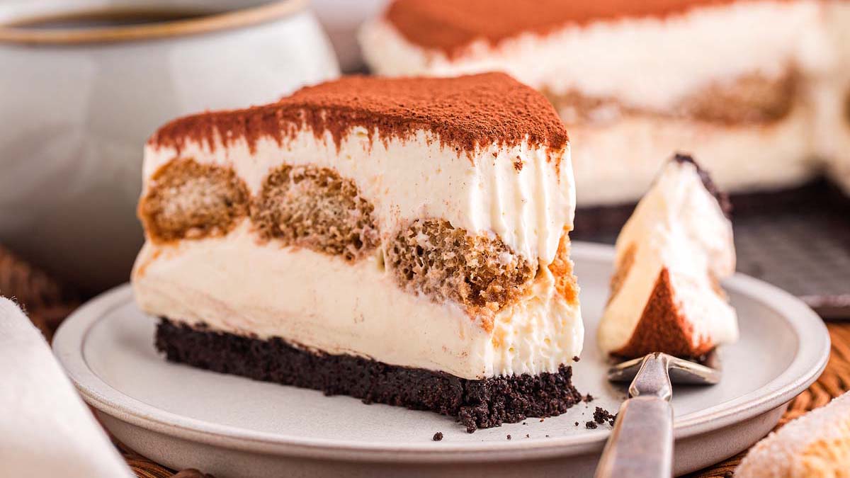 A slice of tiramisu with layers of cream and soaked ladyfingers sits on a plate with a fork, topped with cocoa powder.