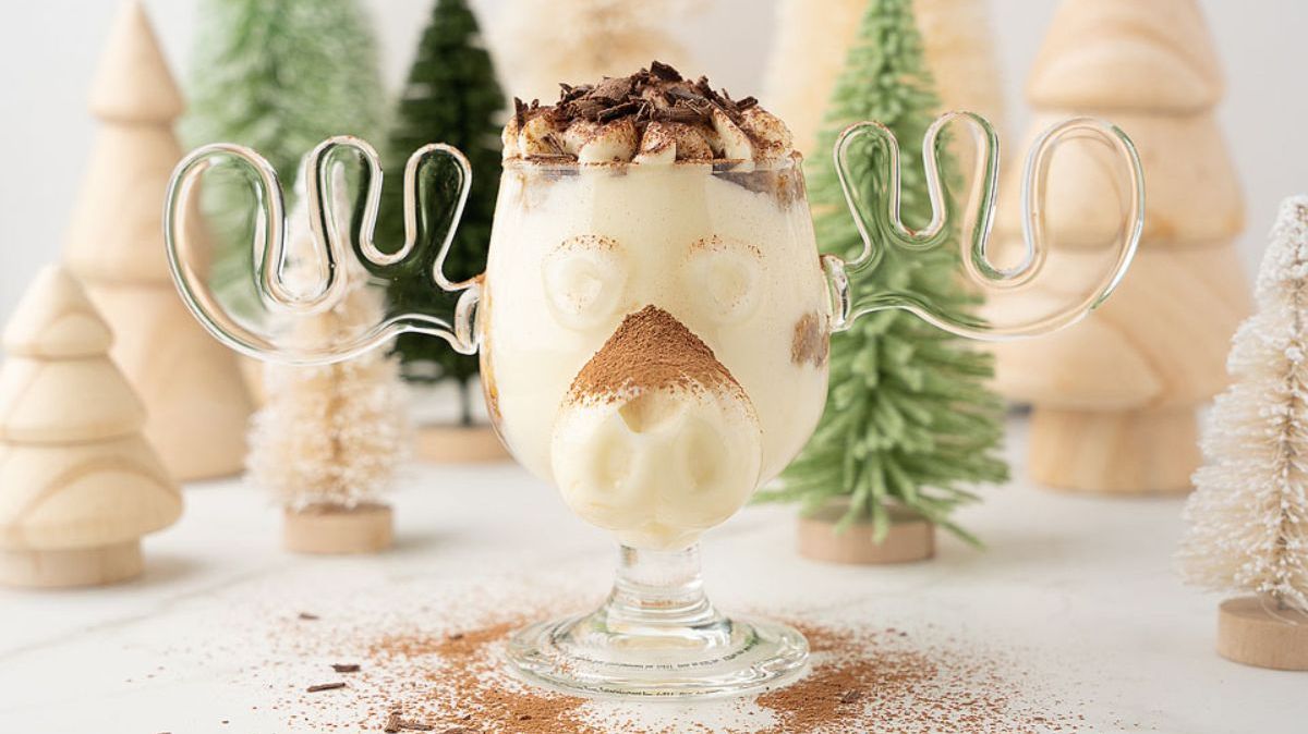 A glass shaped like a moose head filled with eggnog, topped with whipped cream and chocolate shavings, sits on a white surface with wooden and felt Christmas trees in the background.