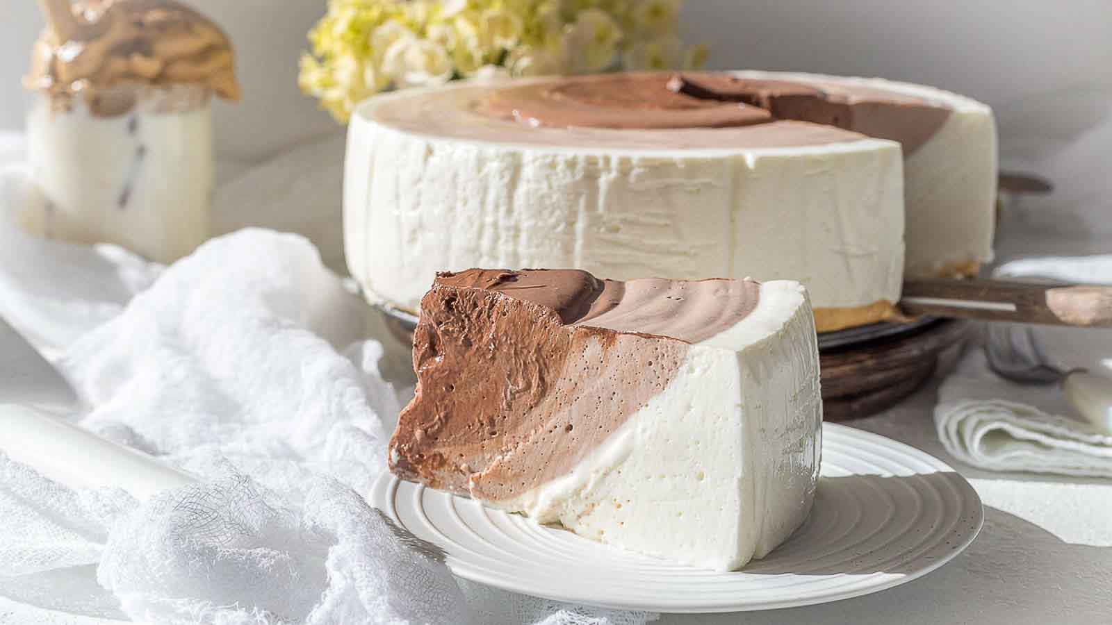A slice of layered chocolate and vanilla mousse cake sits on a white plate, with the remaining cake and a jar in the background.