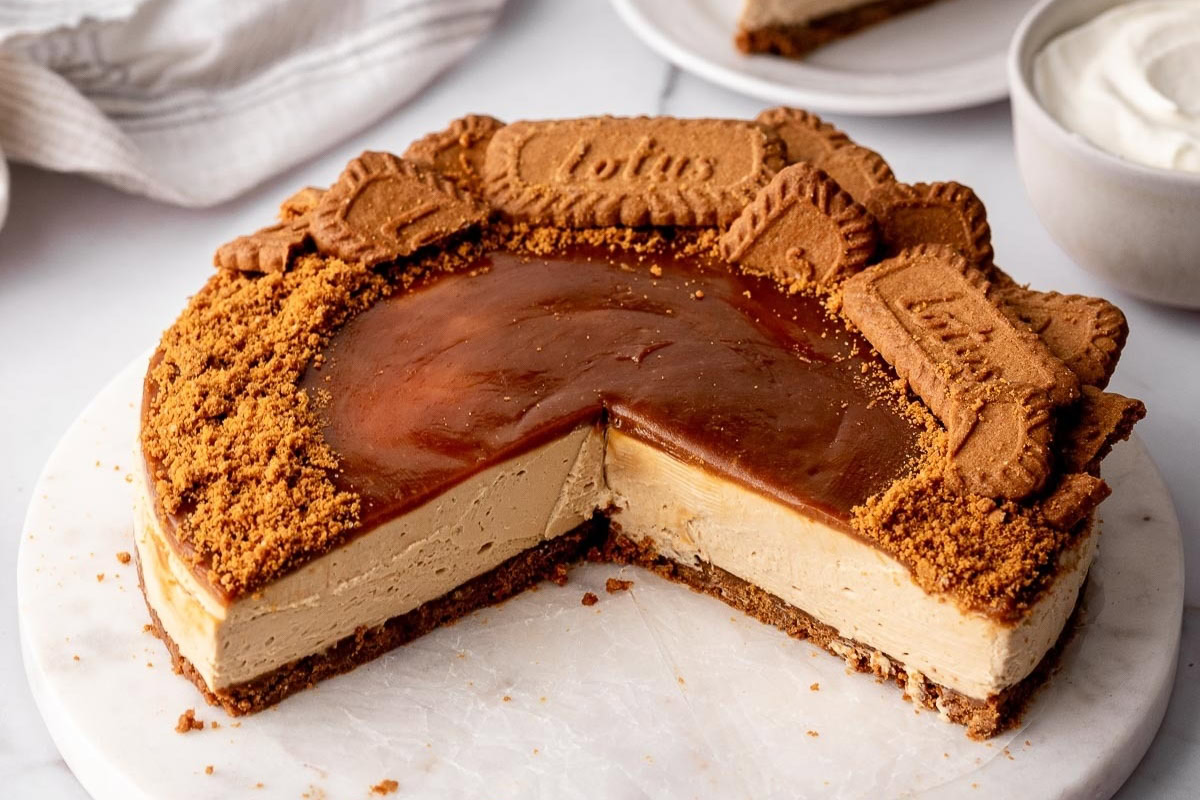 A cheesecake with a caramel topping, decorated with whole and crushed Lotus Biscoff cookies, sits on a marble surface with a slice removed.