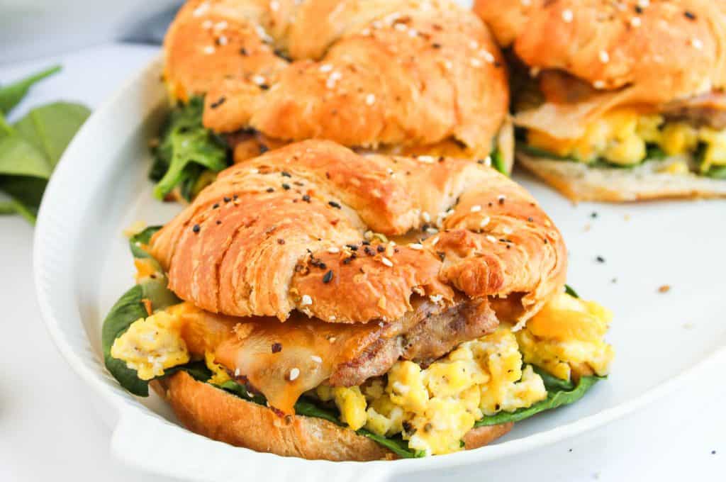 A croissant sandwich filled with scrambled eggs, sausage, cheese, and spinach sits on a white plate, with another similar sandwich in the background.