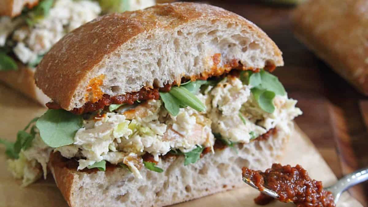A close-up of a sandwich with shredded chicken, arugula, and a spread on crusty bread, with a spoonful of reddish spread nearby.