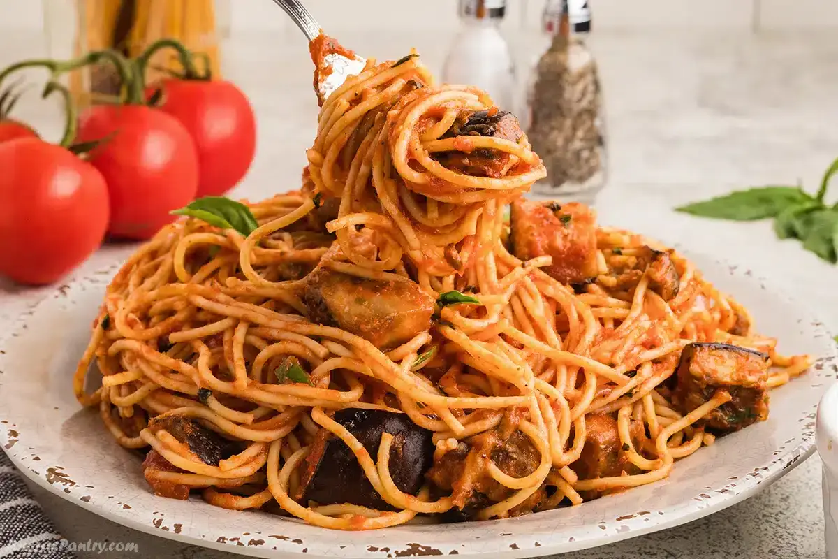 A plate of spaghetti with tomato sauce, eggplant chunks, and herbs, with a fork lifting a bite; fresh tomatoes and seasonings in the background.