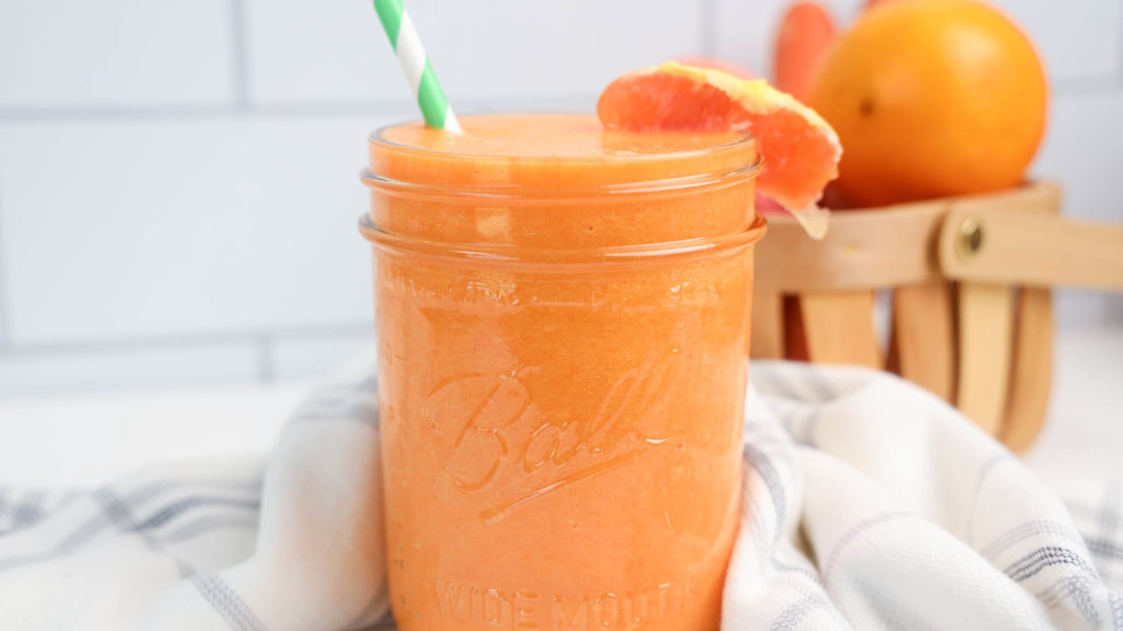 A mason jar filled with orange smoothie, garnished with a grapefruit slice and a striped straw, sits on a cloth with whole grapefruits in a basket in the background.