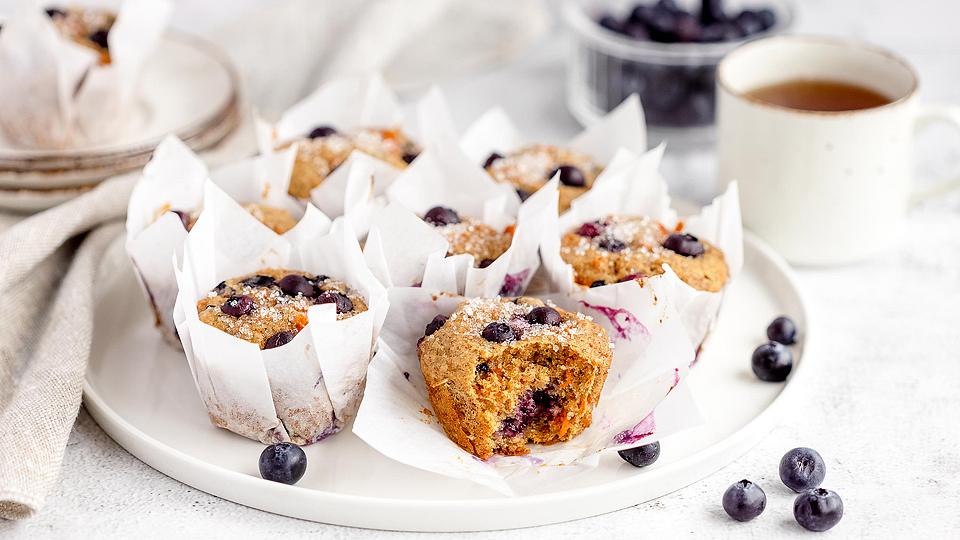 A plate of blueberry muffins in white paper wrappers sits on a table next to fresh blueberries, a cup of tea, and stacked plates.