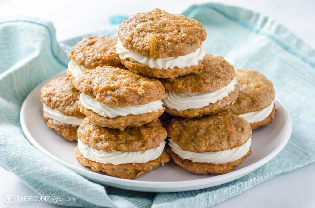 A plate of carrot cake sandwich cookies with cream cheese filling stacked on a white plate, placed on a light blue cloth.