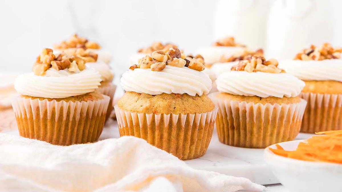 Six cupcakes with white frosting and chopped walnuts on top are arranged on a white surface with a white cloth and a partial bowl visible.