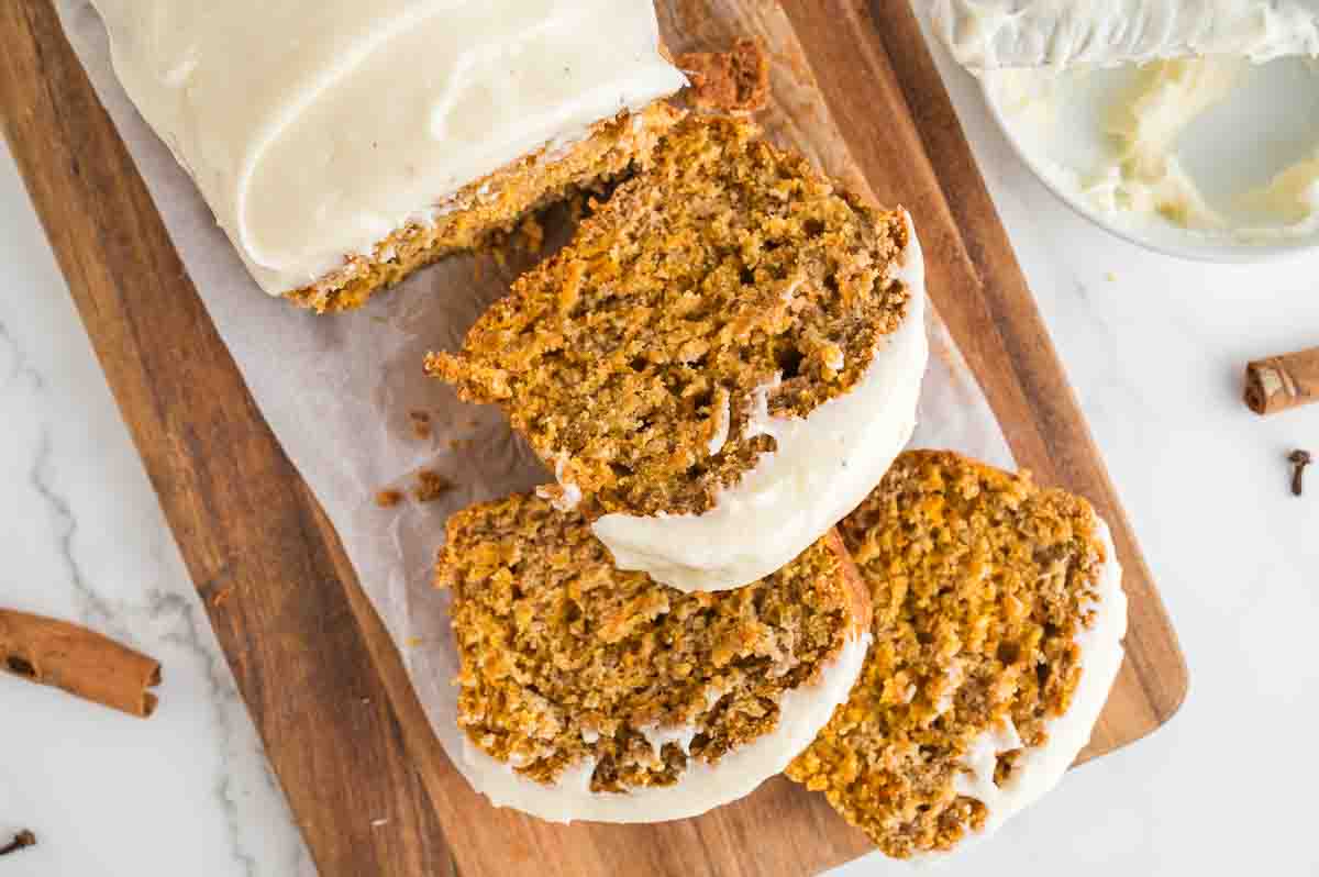 A loaf of carrot bread with white icing is sliced on a wooden cutting board, with three slices arranged in front of the loaf.