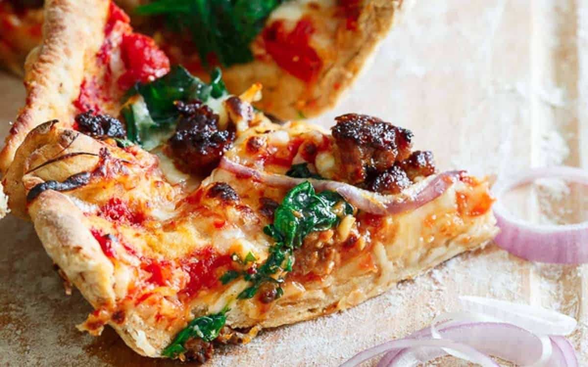 A slice of deep-dish pizza topped with sausage, spinach, red onions, tomato sauce, and melted cheese sits on a wooden surface.