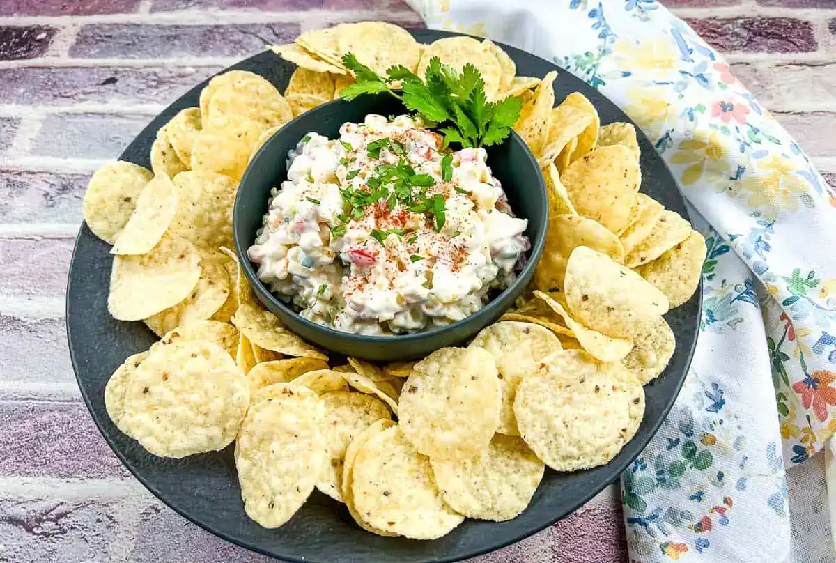 A plate of round tortilla chips arranged around a bowl of creamy dip topped with chopped herbs and paprika, placed on a stone surface with a floral napkin nearby.