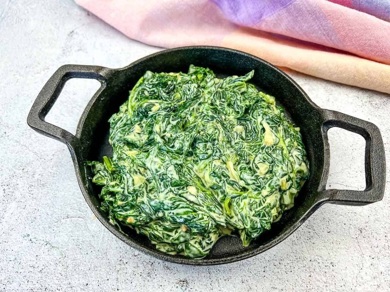 A cast iron dish filled with creamy creamed spinach sits on a light surface near a pastel-colored cloth.