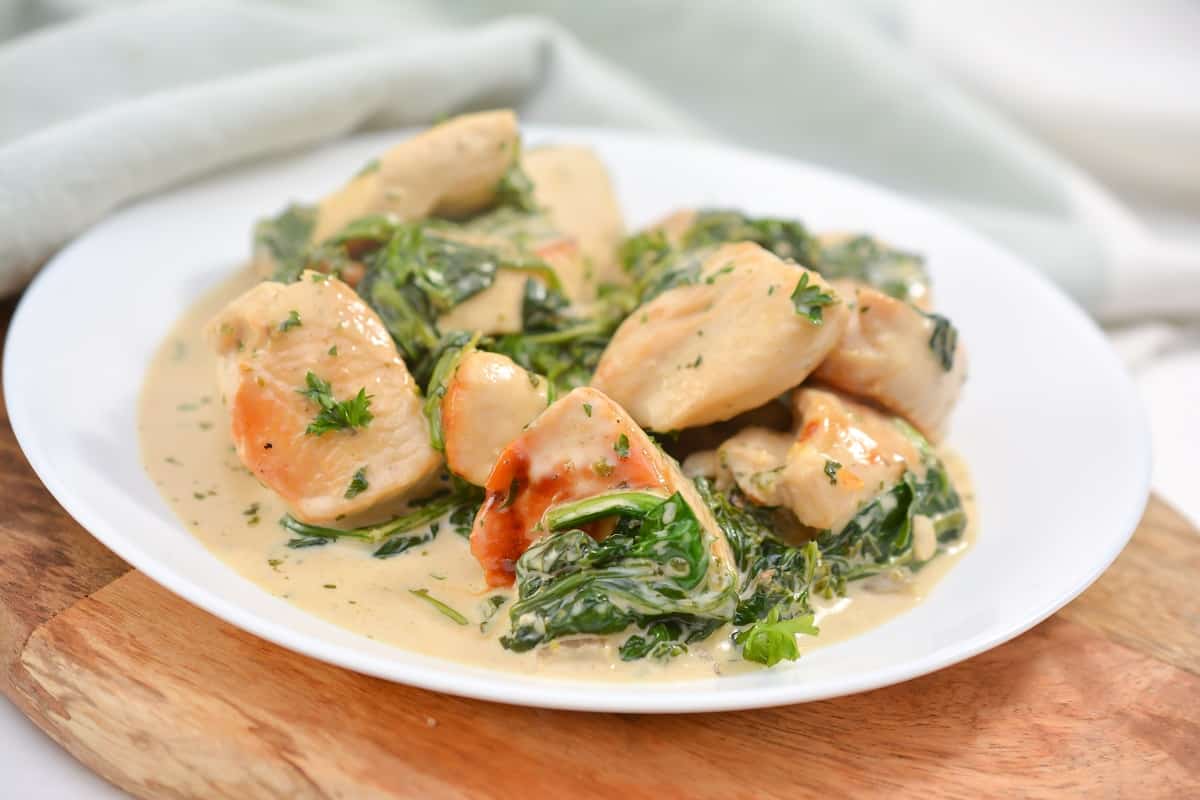 Chicken breast pieces with wilted spinach and tomato in a creamy sauce are served on a white plate atop a wooden surface.