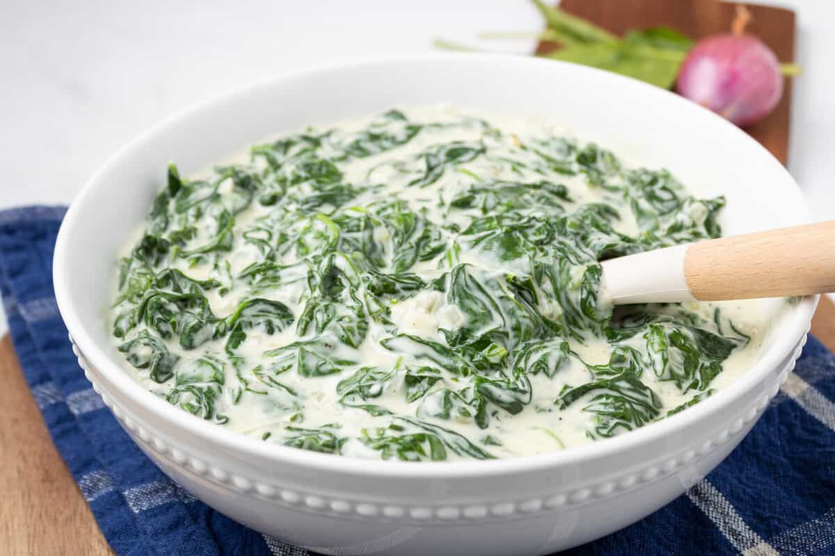 A white bowl filled with creamy spinach dip, with a wooden-handled spoon resting inside, placed on a blue cloth napkin.
