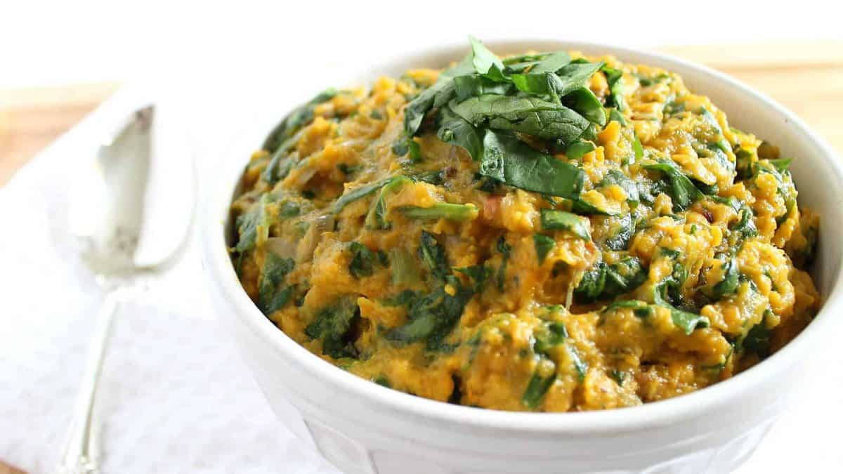 A white bowl filled with a creamy spinach and lentil curry, garnished with fresh spinach leaves, sits beside a spoon on a white cloth.