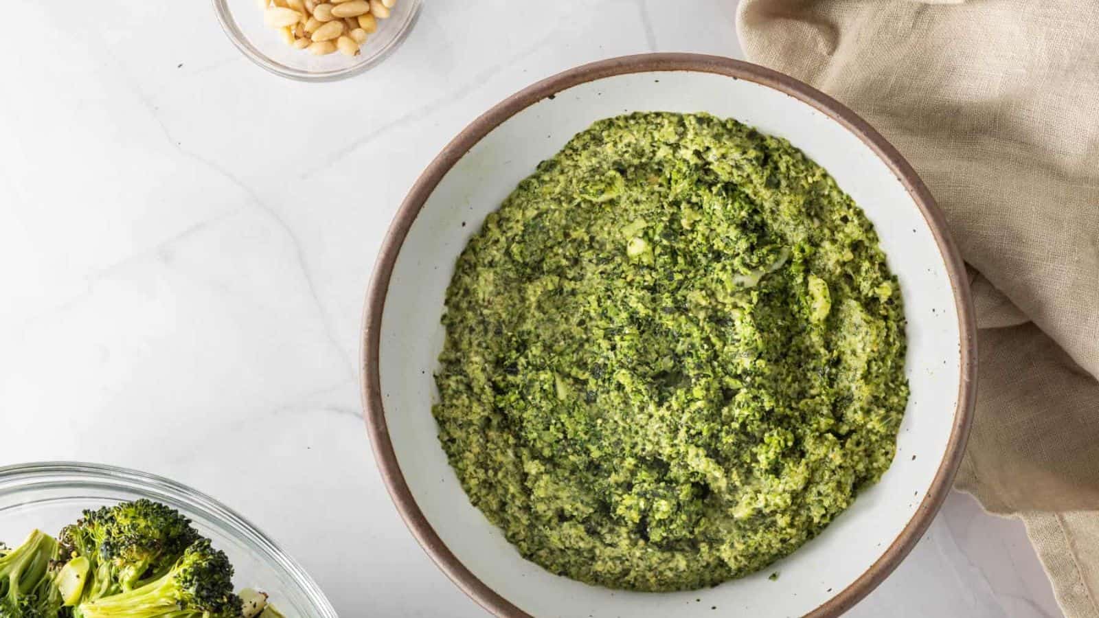 A bowl of green broccoli pesto sits on a white surface next to a napkin, with a small bowl of pine nuts and a dish of cooked broccoli nearby.
