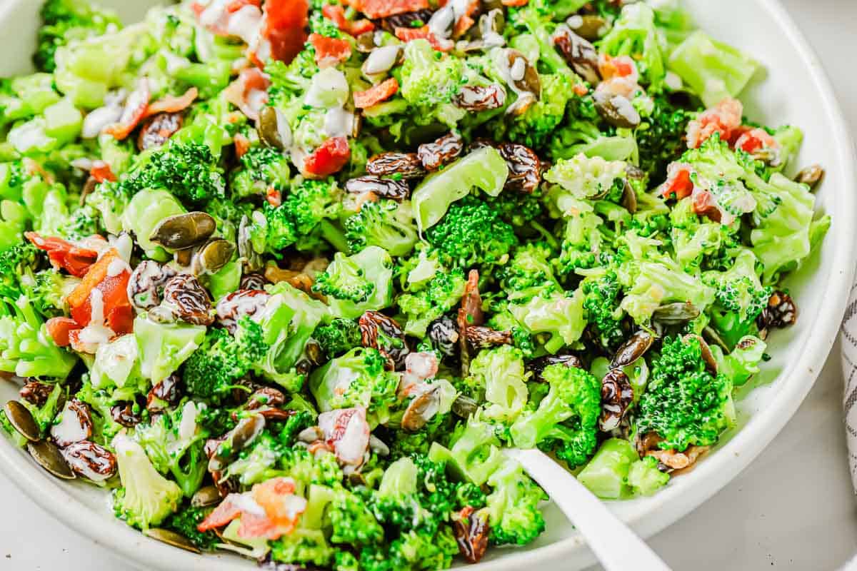 A bowl of broccoli salad with sunflower seeds, raisins, chopped bacon, and a creamy dressing, served on a white plate.