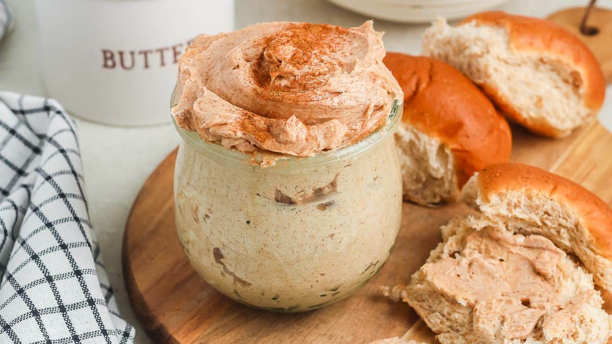 A jar of cinnamon honey butter with a swirl on top sits on a wooden board next to split and spread dinner rolls.