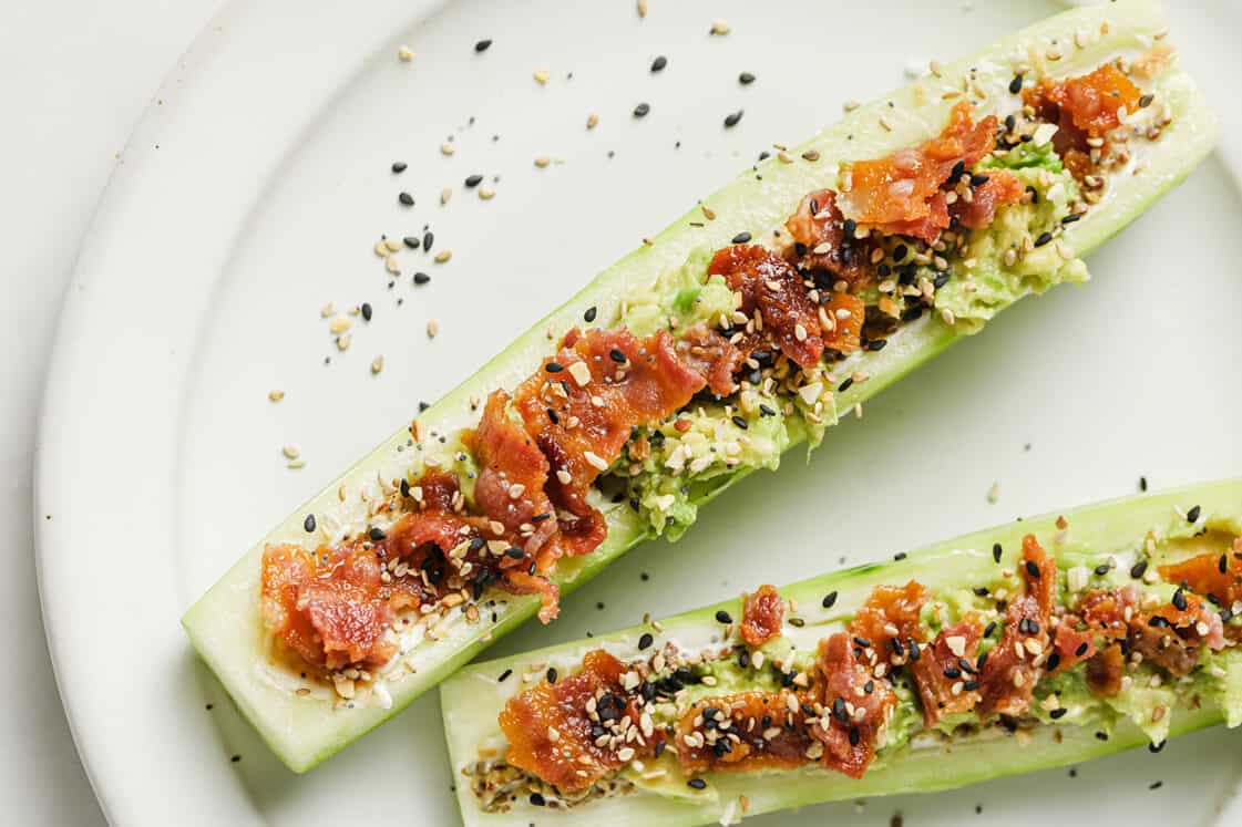 Two cucumber halves filled with mashed avocado, crumbled bacon, and sprinkled with mixed seeds, served on a white plate.