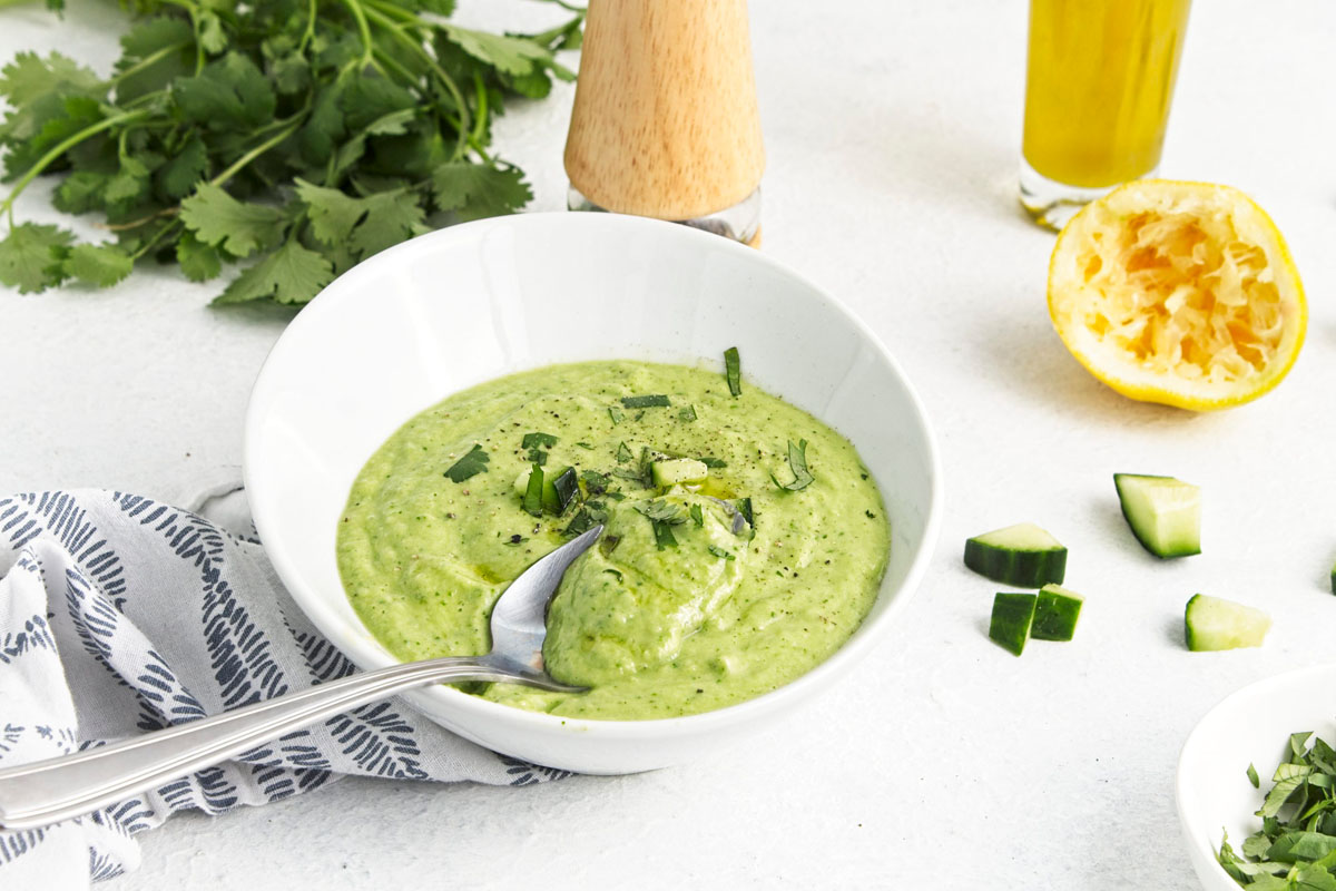 A bowl of green soup garnished with herbs sits on a table with a spoon, surrounded by cucumber pieces, cilantro, a lemon half, and a bottle of olive oil.