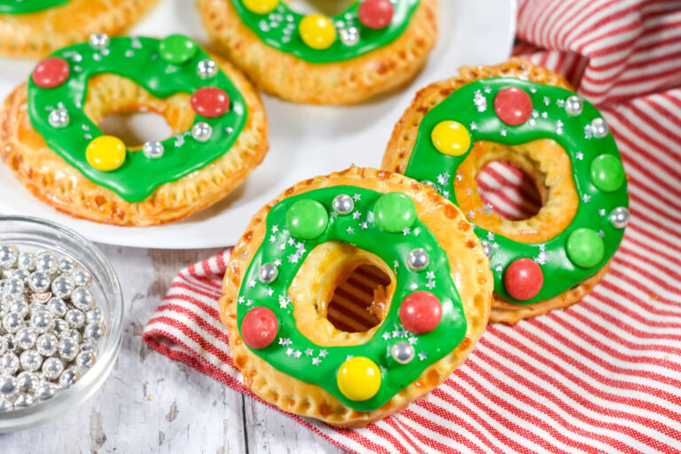 Round pastries decorated with green icing, colored candies, and silver sprinkles, arranged on a white plate and red-striped cloth.