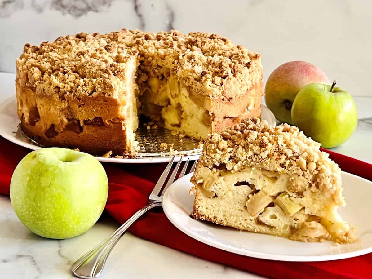 A sliced apple crumb cake sits on a white plate, with a larger cake in the background and whole apples nearby on a red cloth.