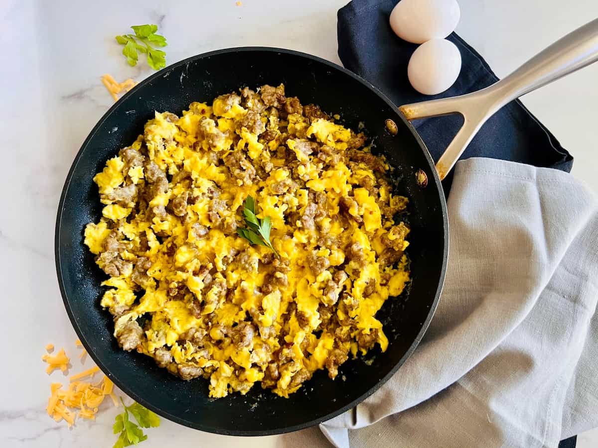 A skillet filled with scrambled eggs and sausage sits on a gray napkin, with two eggs, shredded cheese, and fresh herbs nearby.