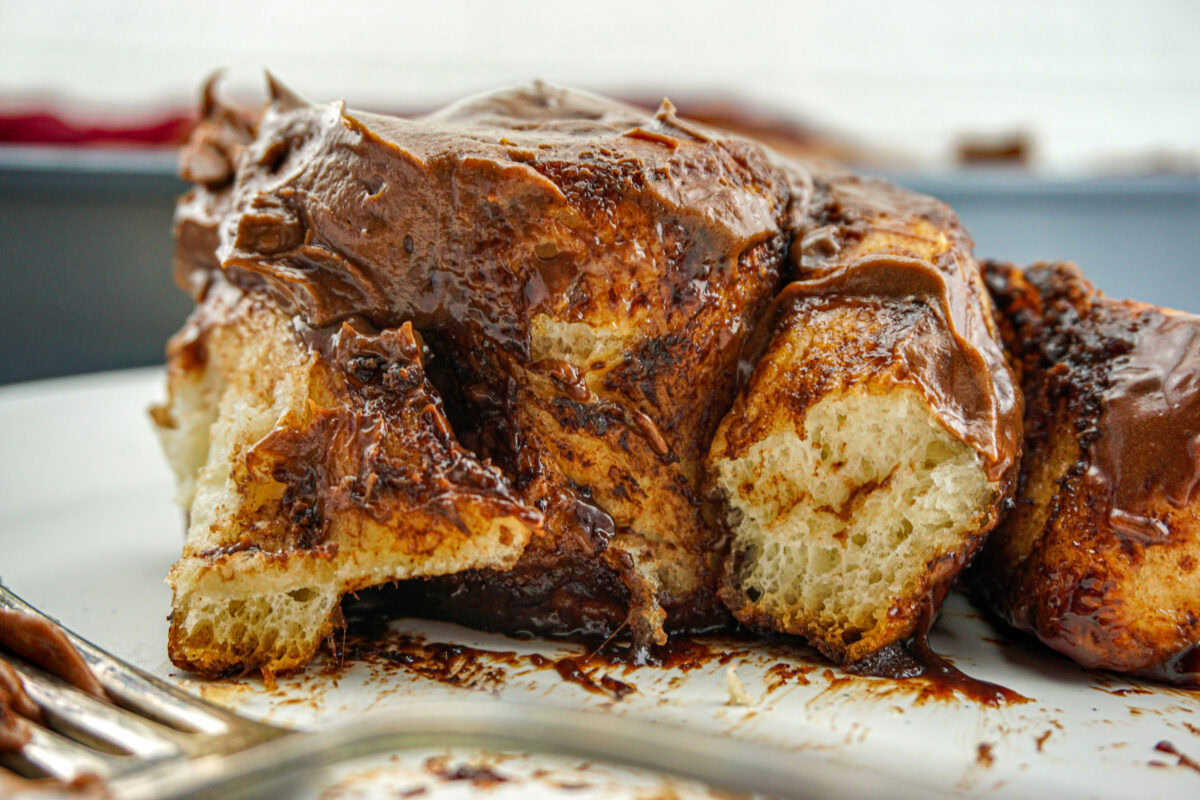 A close-up of a cinnamon roll covered in chocolate frosting on a white plate, with a fork in the foreground.