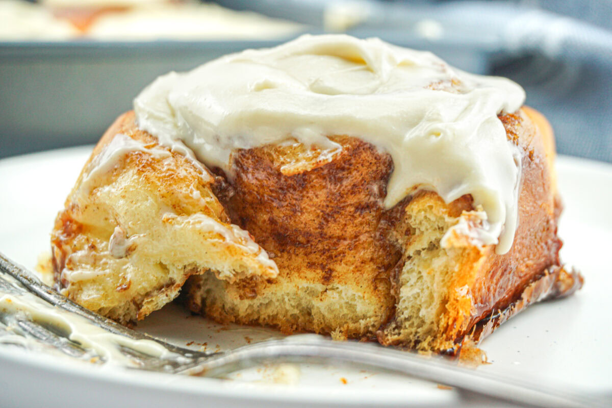 A cinnamon roll with cream cheese frosting sits on a white plate, partially eaten, with a fork resting nearby.