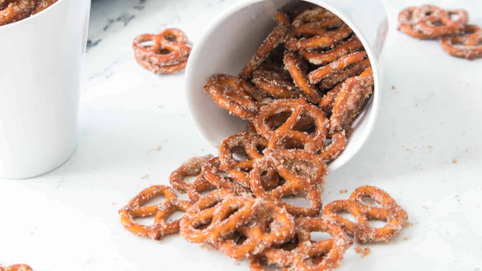 A white cup tipped over, spilling sugared pretzels onto a white surface. More sugared pretzels are scattered around the cup.