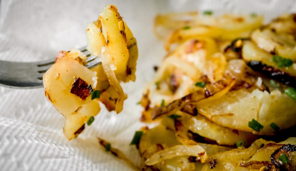 A fork holds up cooked sliced potatoes and onions, garnished with chives, with more pieces visible on a napkin in the background.