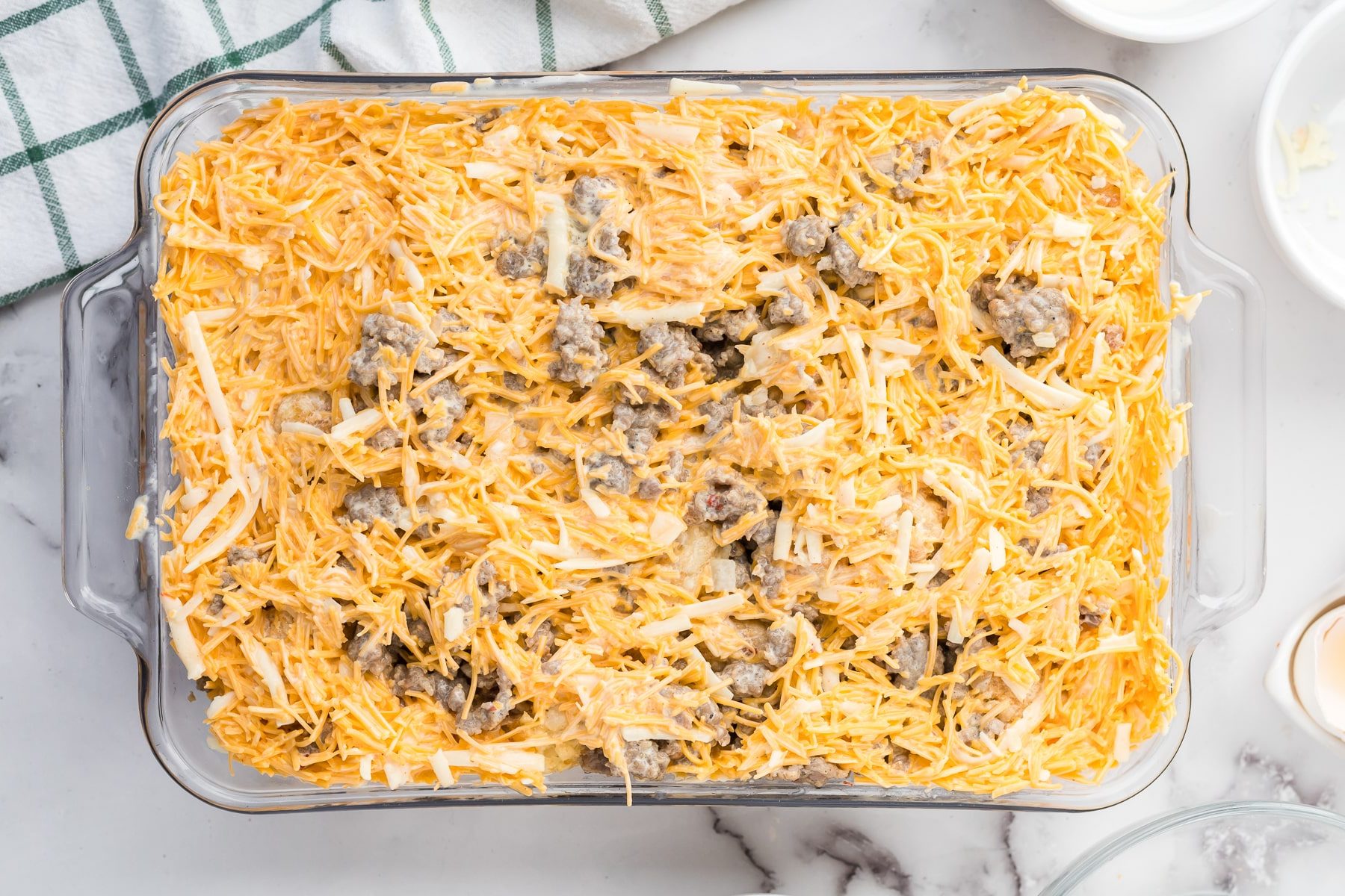 A glass baking dish filled with an unbaked mixture of shredded cheese, cooked sausage, and other ingredients on a marble countertop.